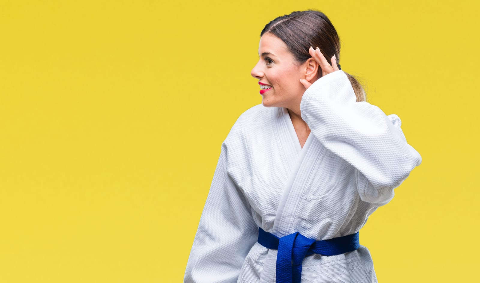Woman wearing karate kimono uniform over isolated background smiling with hand over ear listening