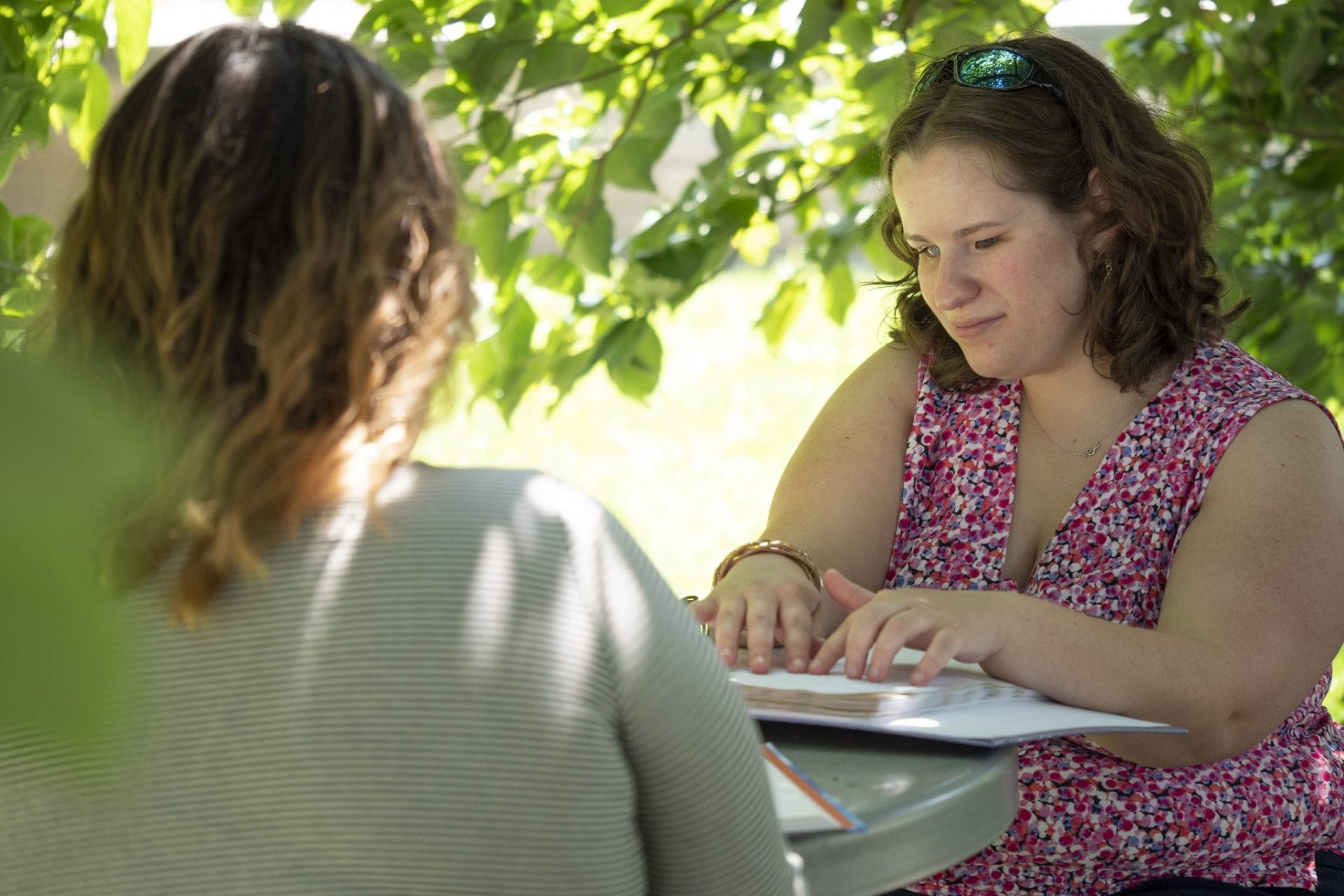 A student reads a braille textbook while sitting outside under a tree with a friend