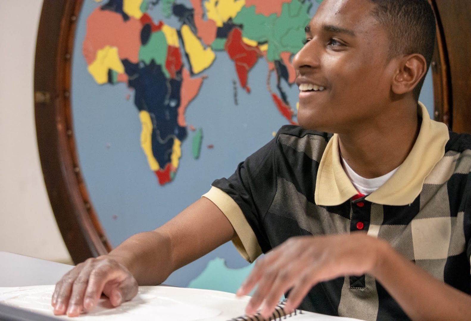 Teenage boy reading braille and smiling in front of a tactile map of the world