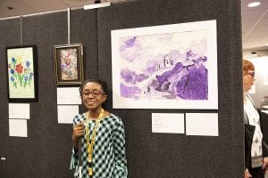 a young artist, smiling and holding her white cane, stands in front of her purple landscape artwork