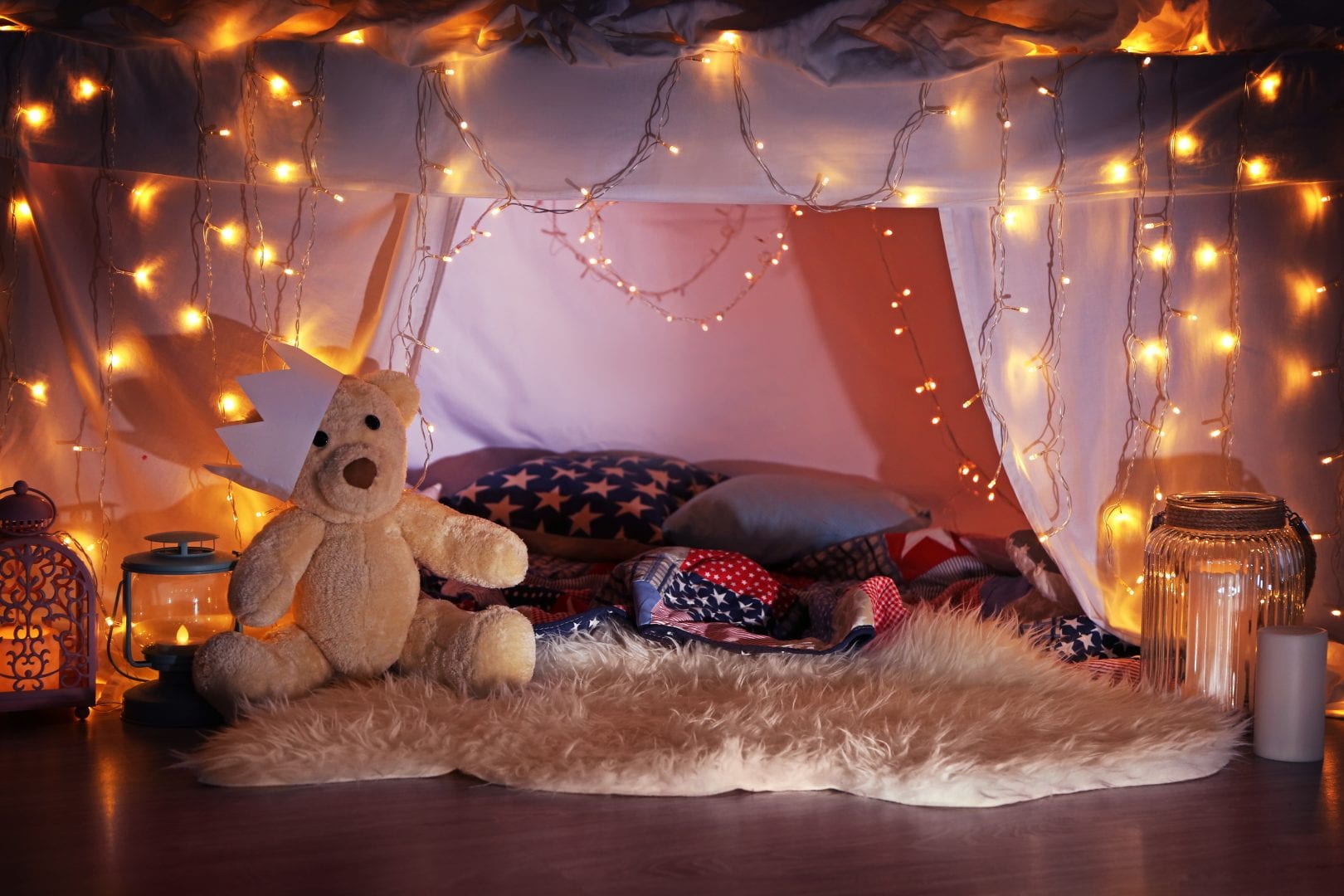 an indoor tent built out of sheets featuring, pillows, blankets, a teddy bear, and twinkle lights.