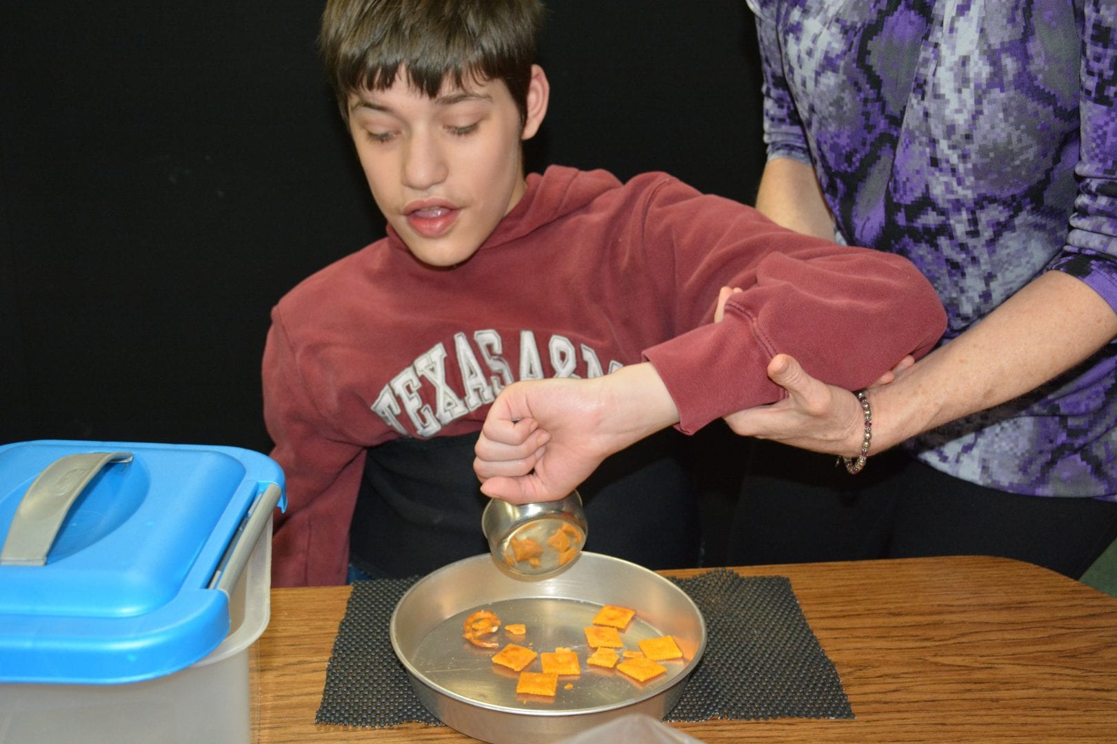 A teacher provides elbow support as a learner uses a measuring cup to pour his snack into a pan