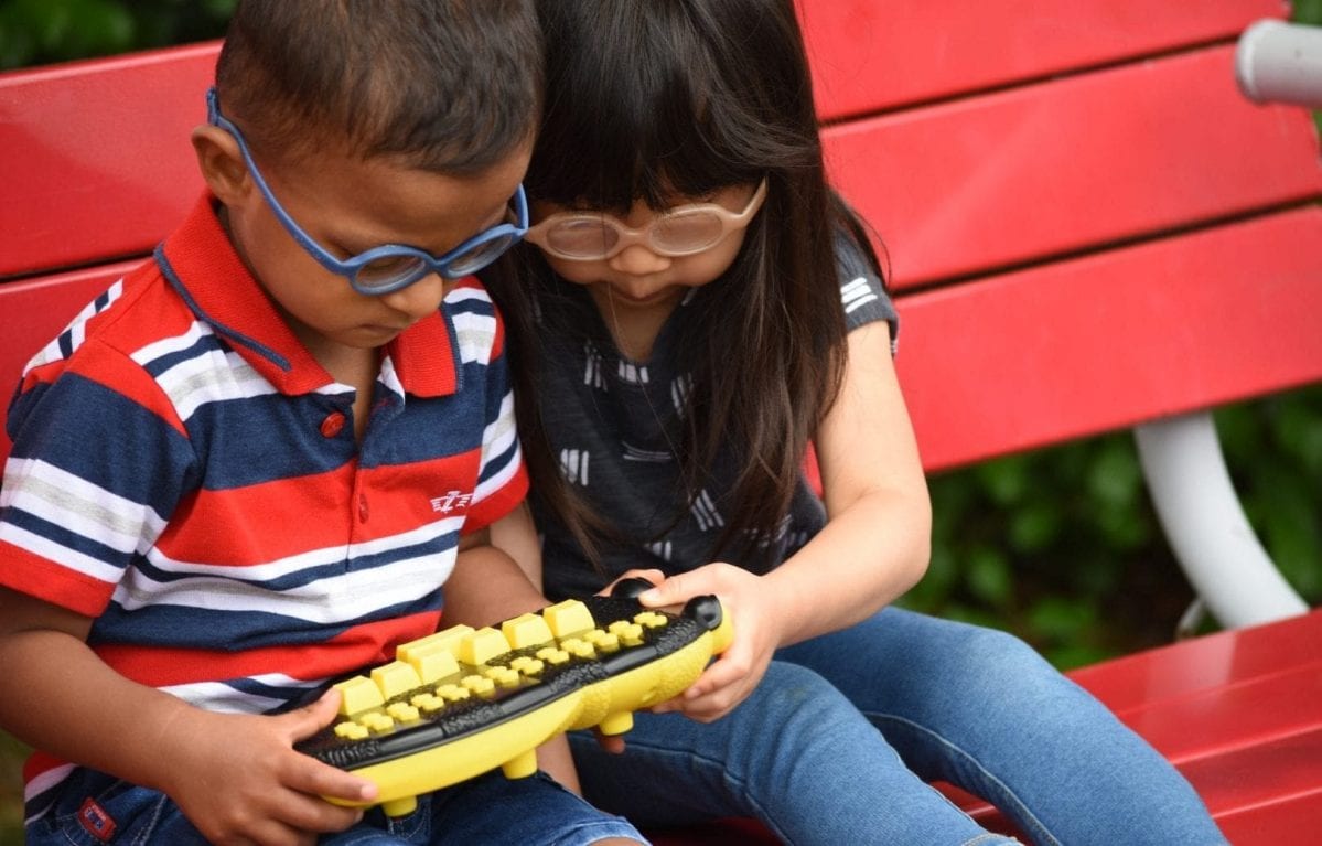 a little boy and girl, both wearing glasses, sit on a red bench and play with Braille Buzz together.