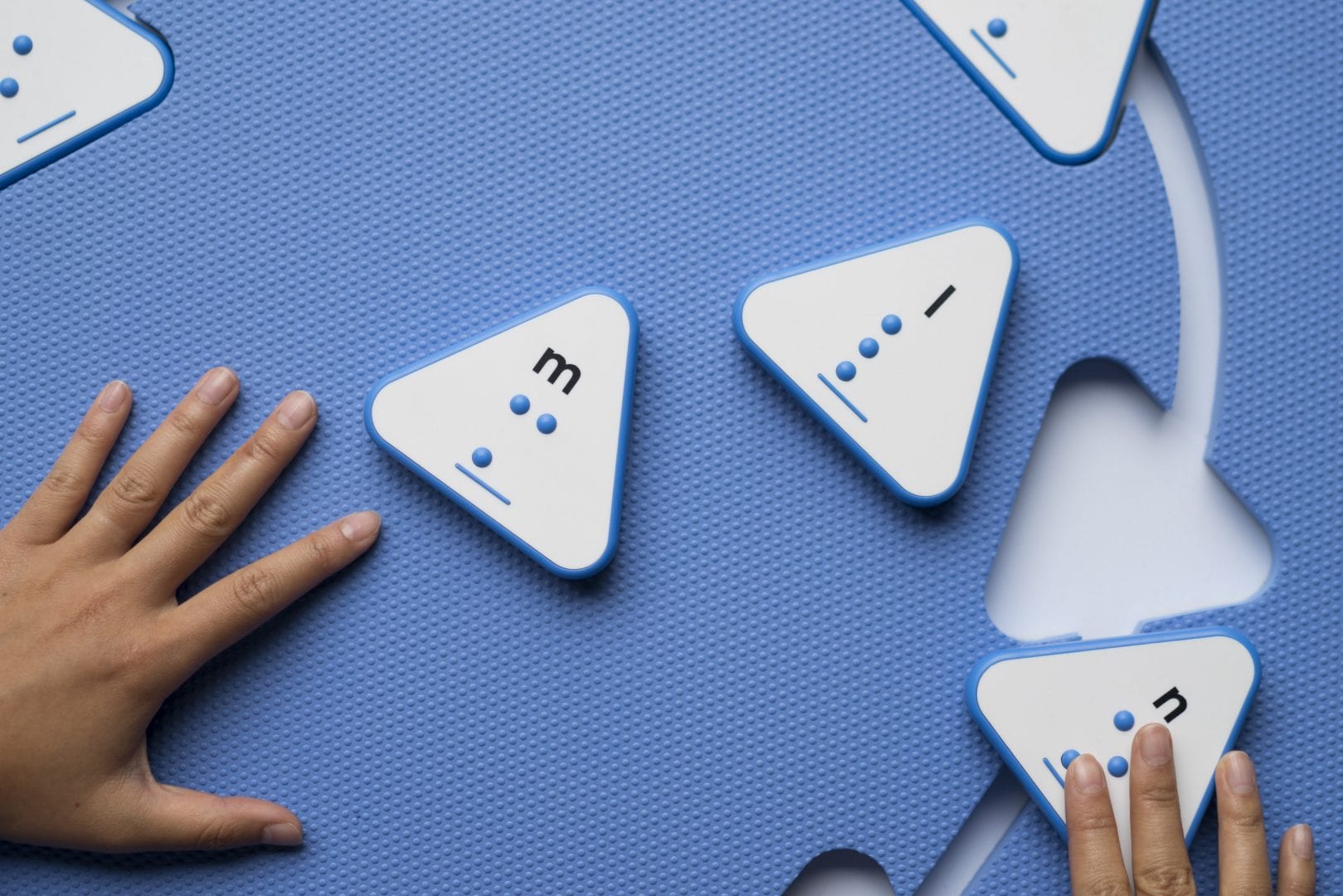 Two hands placing triangle print-braille tiles into the cutouts of the blue Reach & Match mat.