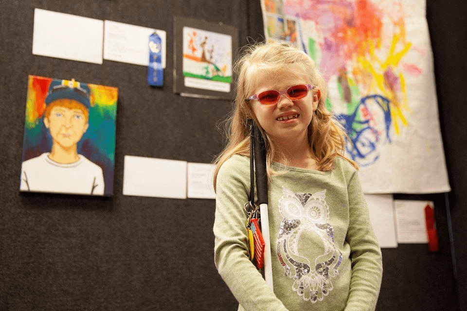 Young girl in pink sunglasses holding a white cane while standing in front of a wall of art