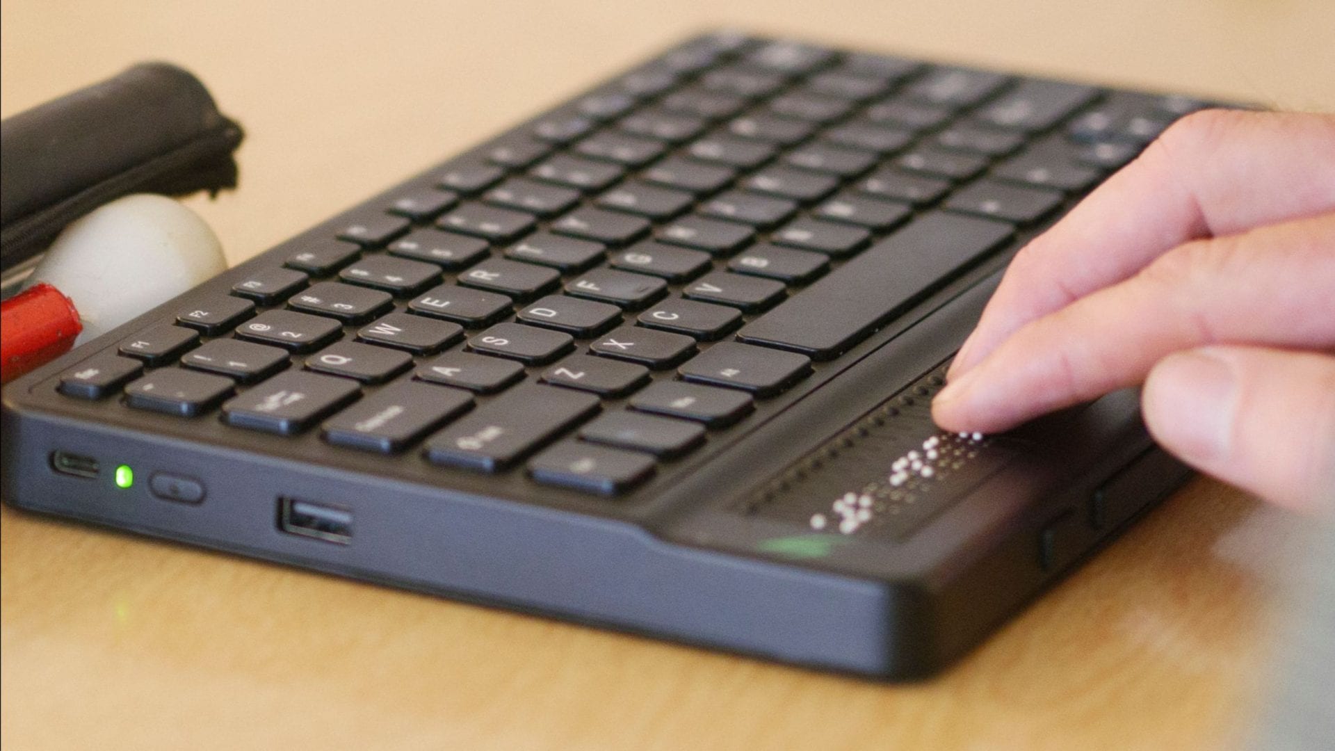 A person's hand reading the 40 cell braille display on the Mantis Q40. A folded up white cane sits on the table.