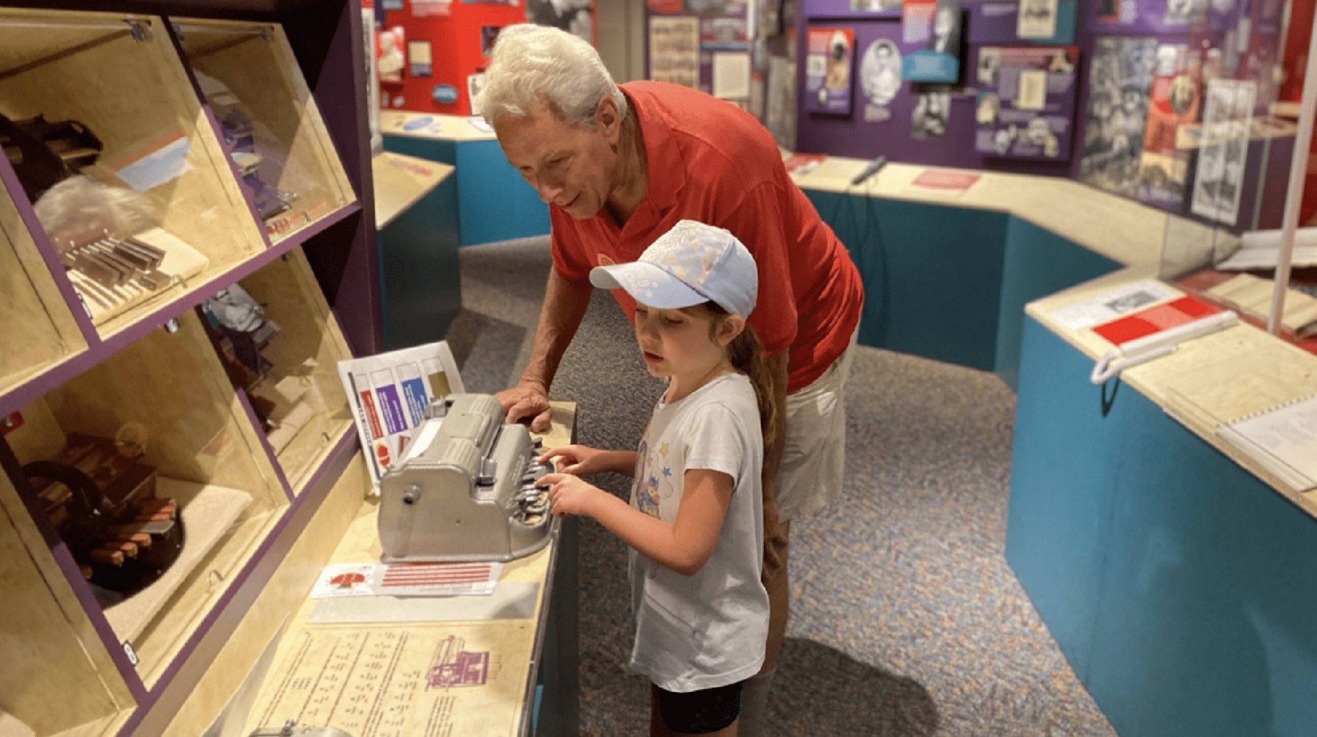 An older man watches intently as a young girl wearing a baseball hat demonstrates how to use the braillewriter.