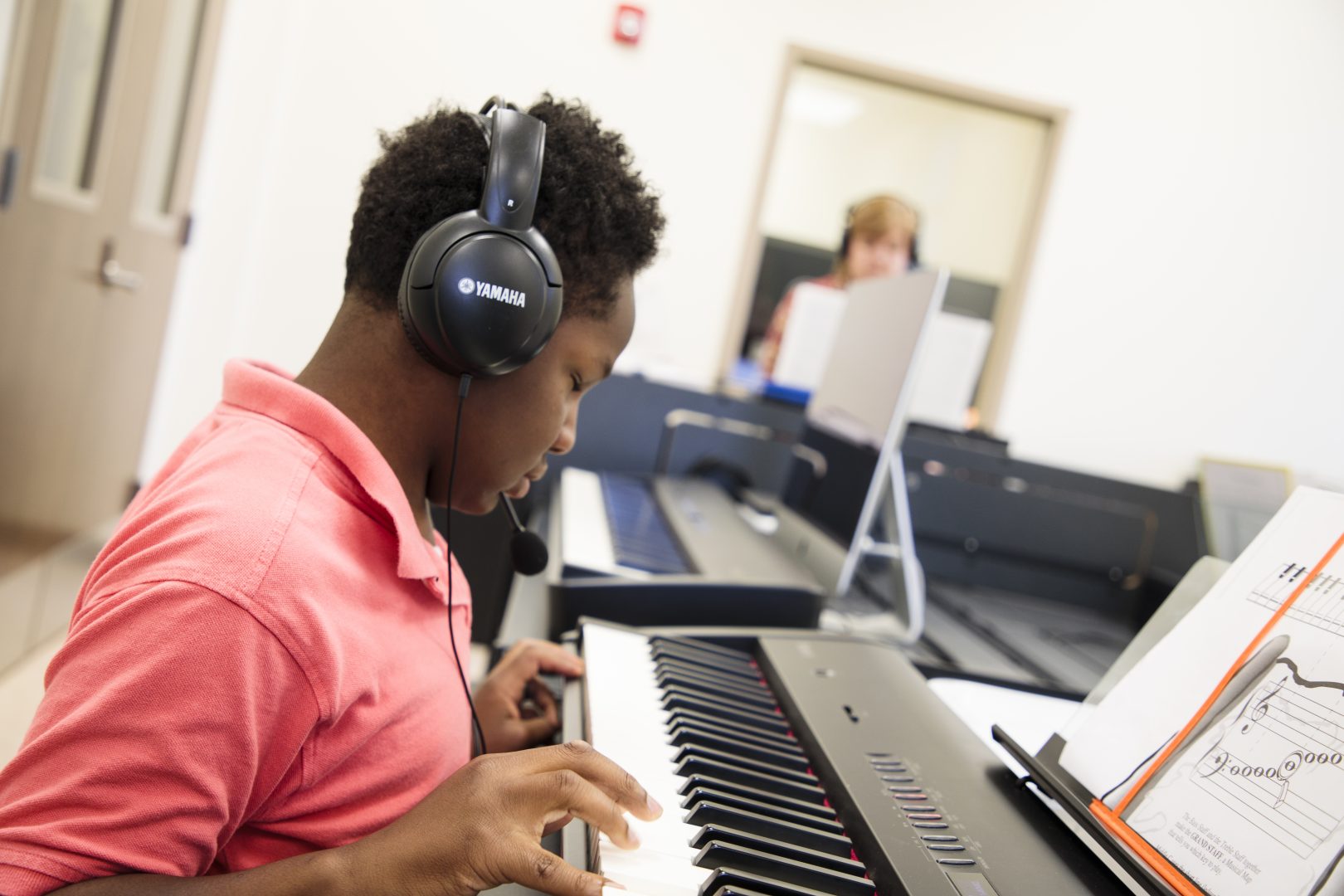 An elementary-age student playing the keyboard and wearing headphones. A teacher can be seen in the background.