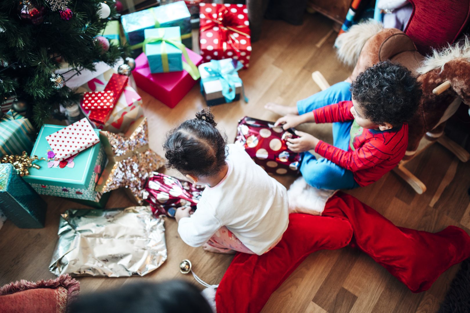 birds eye view of two children sitting on the floor opening colorful presents in front of a christmas tree
