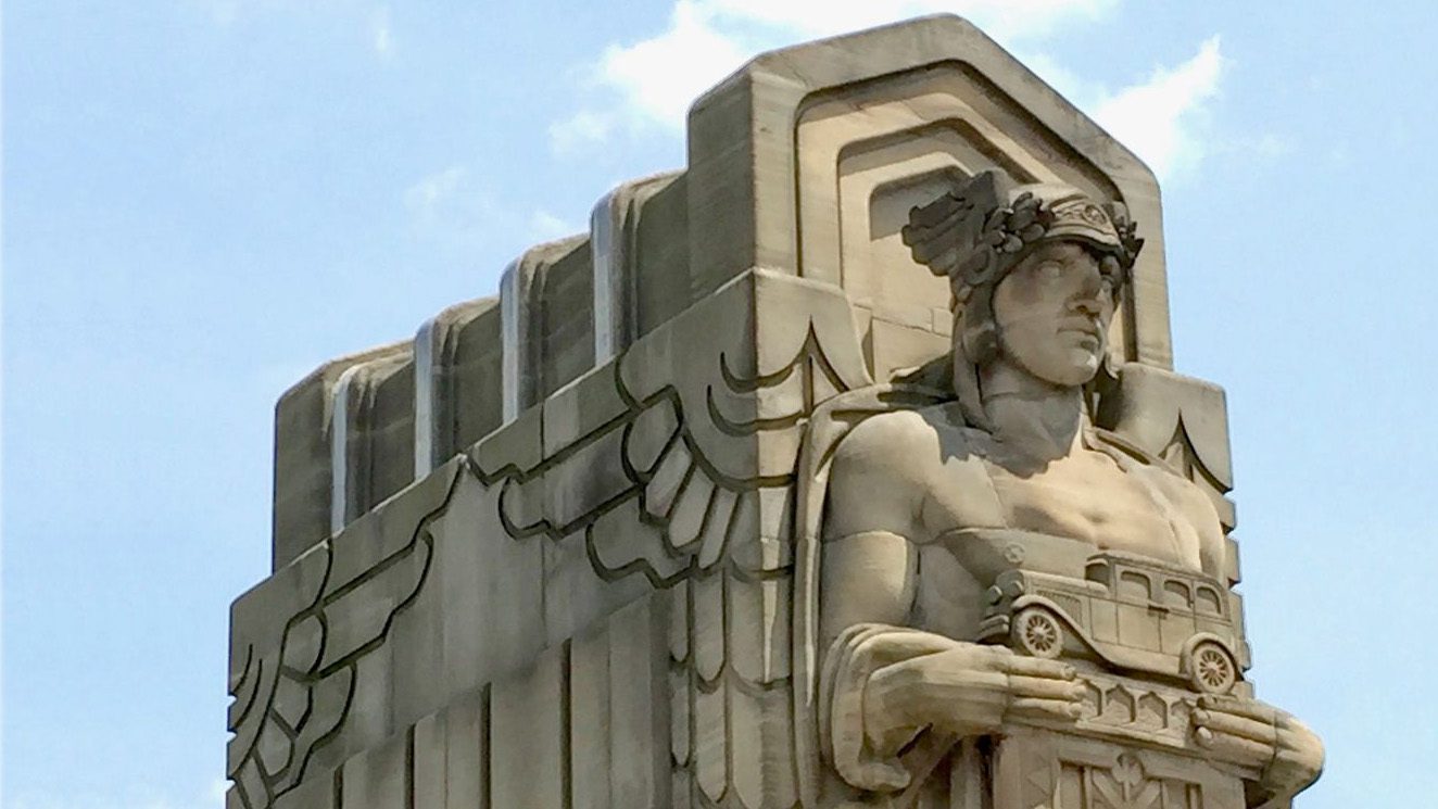 Statue of a man in front of a wall. He is wearing a crown of leaves, with wings at either side, and holding a model of a 1930s automobile.