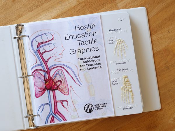 Health Education Tactile Graphics binder opened to show the cover of the Instructional Guidebook for Teachers and Students. Part of the first tactile graphic showing “hand detail” and “foot detail” is also shown. The carpal bones and phalanges are listed in braille for the “hand detail,” while the tarsal bones and phalanges are listed in braille for the “foot detail.”