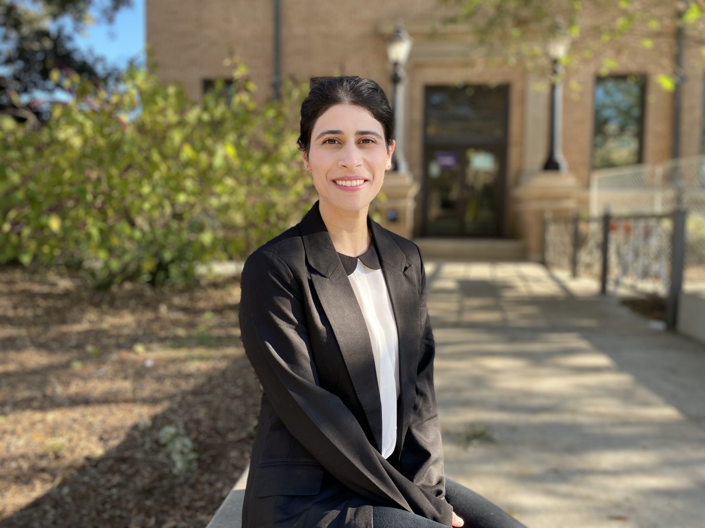 Nabiha sitting outside in front of a landscaped building entrance. She is smiling and wearing a black blazer and white button down shirt.