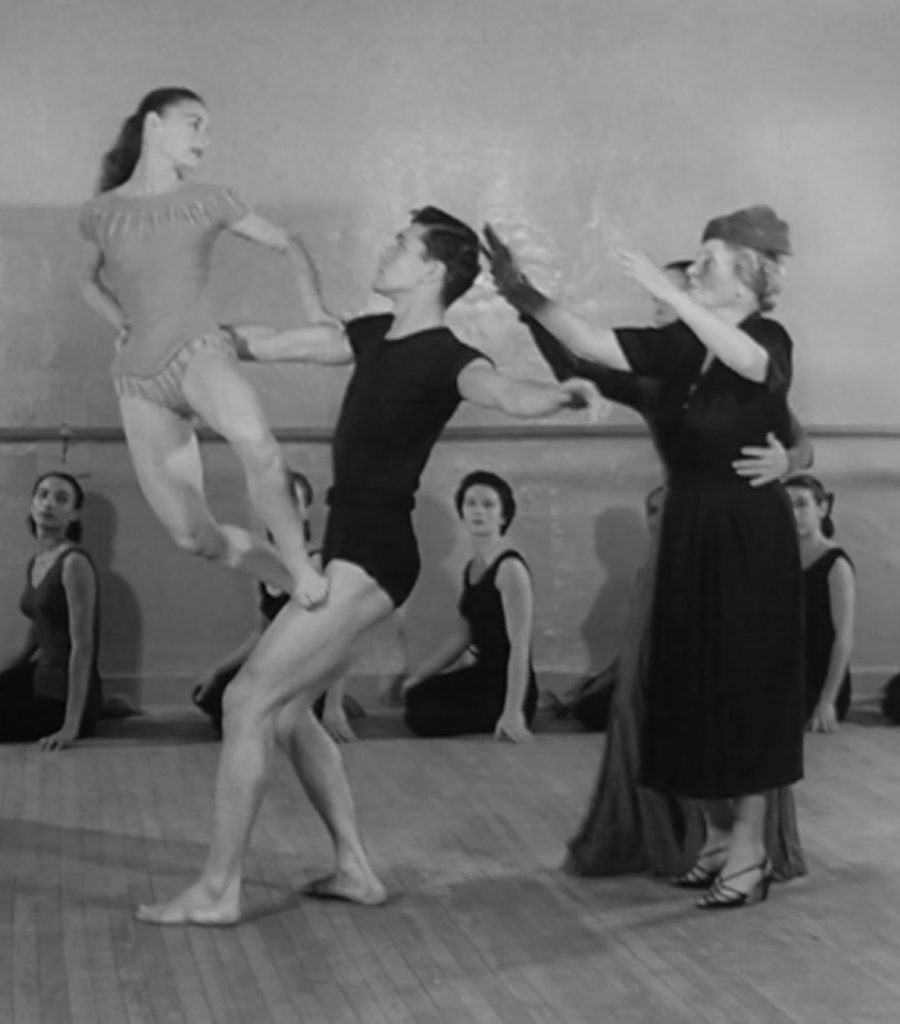 A black and white photo of Helen Keller with her arms upraised in motion with three dancers, one has been lifted in the air. Other dancers sit along a wall with a ballet bar in the background.