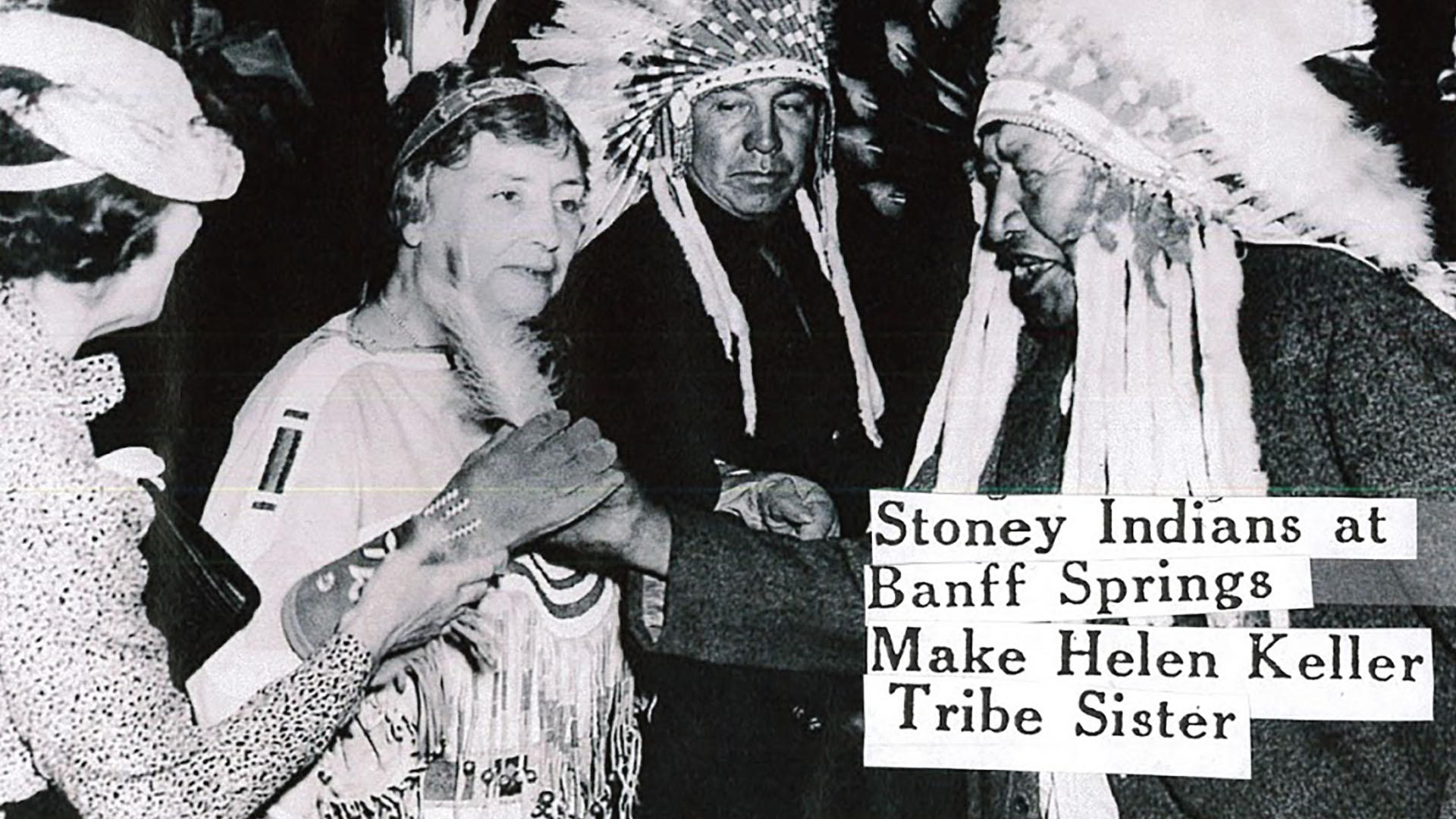 Polly Thomson and Helen Keller stand with two Native American men wearing robes and full feather headdresses. Helen wears a headband with two eagle feathers and a leather dress with fringe and beading. A caption reads, ”Stony Indians at Banff Make Helen Keller Tribe Sister.”