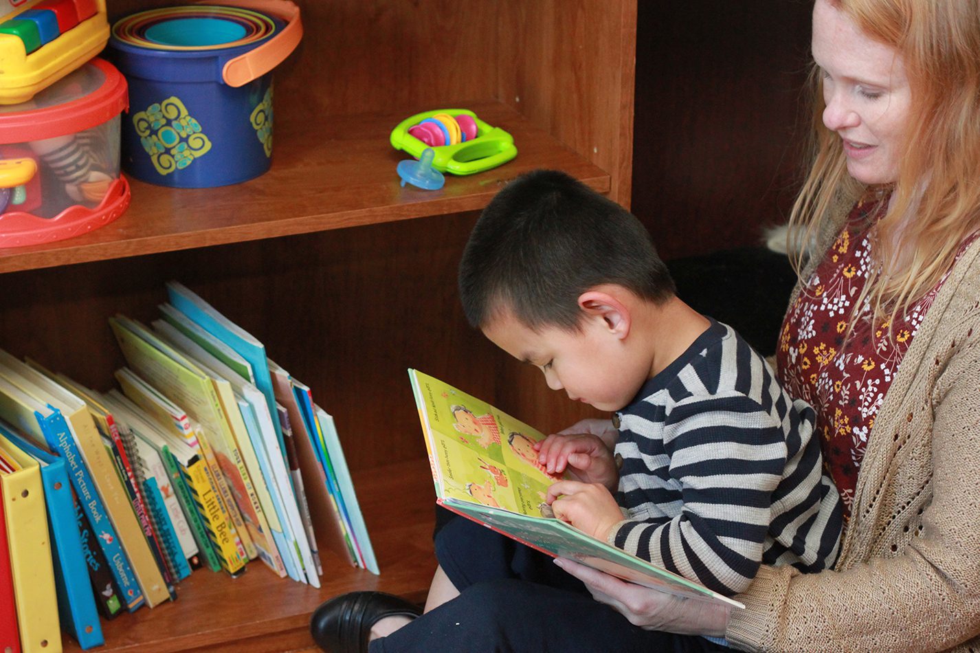 A mother and child sitting on the floor next to a bookcase reading a print/braille book together.
