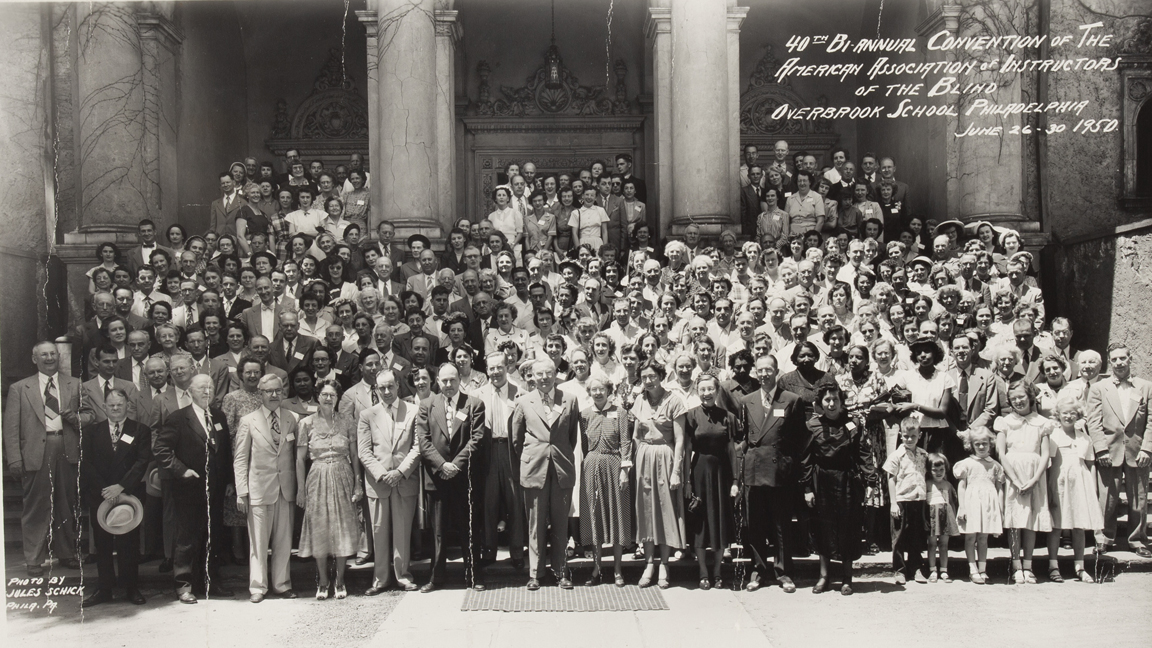 A black and white panorama photo of a large group of people dressed in 1950s clothing standing on the steps of a building.
