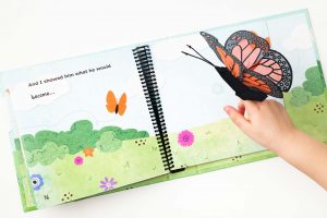 A child’s hand hovers above the page of the book wearing the included orange and black butterfly finger puppet. The pages of the book are illustrated with a green field and flowers and features print and braille text “And I showed him what he would become…”
