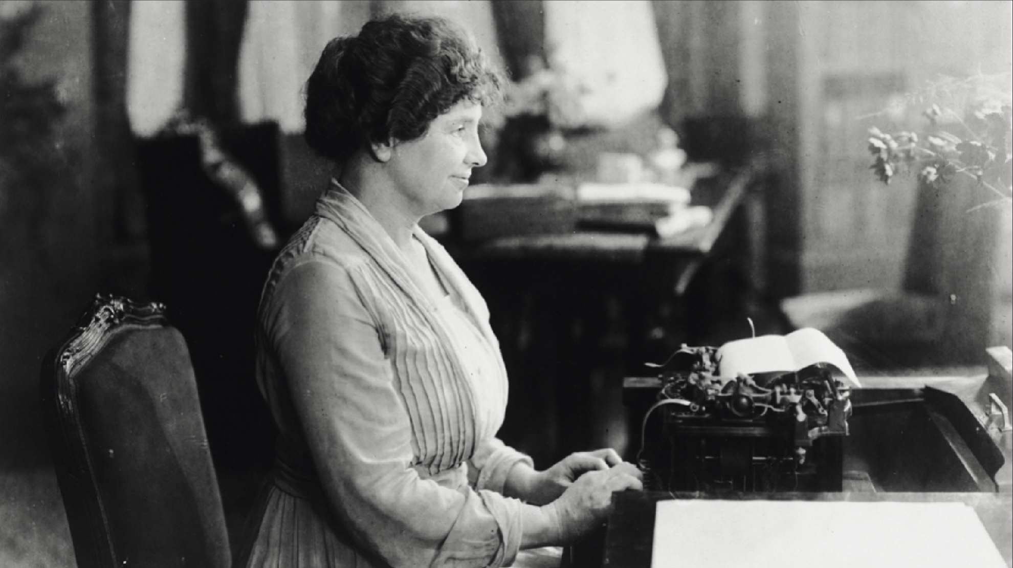 Helen Keller sits at a desk, her fingers poised on the keys of a typewriter. Her hair is piled on top of her head and she wears a light-colored dress with pleating down the front.