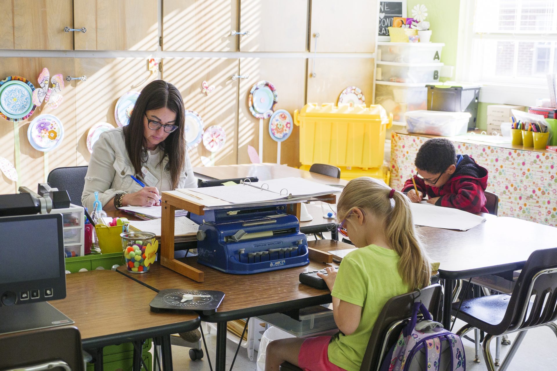 A teacher and two students in a classroom. One student in glasses writes with paper and pencil. The other student in glasses has a Perkins brailler and refreshable braille display in front of them. A tabletop magnifier is sitting off to the side.