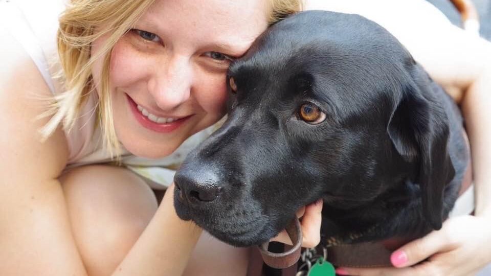 Close up photo of Justine leaning in close and smiling, hugging onto Dove, a black lab.